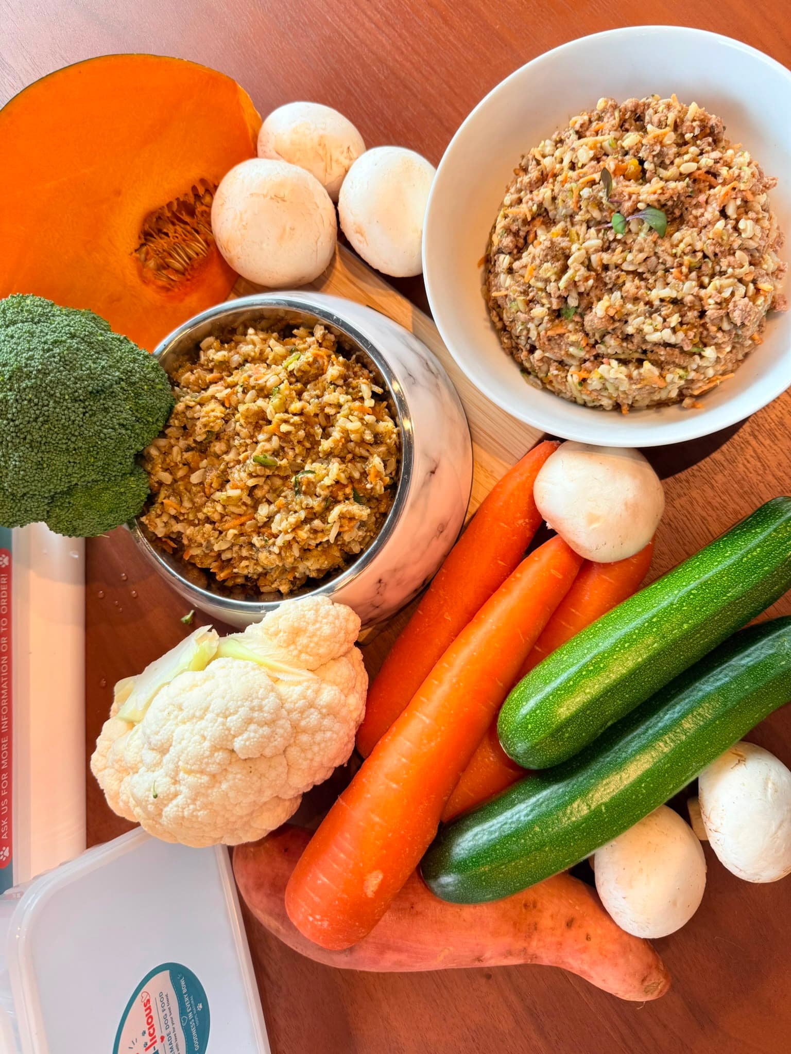 Overhead flatlay of pumpkin, mushrooms, broccoli, cauliflower, carrot and zucchini surrounding bowls of finished Paw-a-licious food